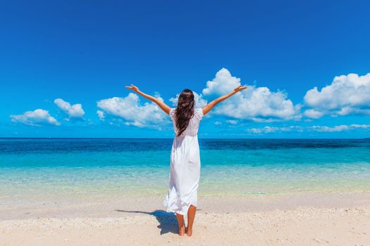 Woman in white dress posing in tropical sea beach with arms raised