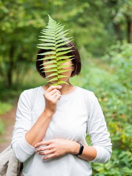 Young woman is hiding her eyes with fern leaf. Symbol of life, tranquility and unity with nature. Summer in forest.