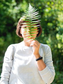 Young woman is hiding her face with fern leaf. Symbol of life, tranquility and unity with nature. Summer in forest.