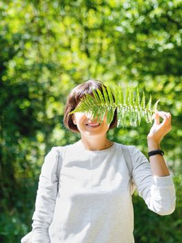 Young woman is hiding her eyes with fern leaf. Symbol of life, tranquility and unity with nature. Summer in forest.