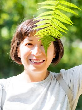 Young woman is hiding her eyes with fern leaf. Symbol of life, tranquility and unity with nature. Summer in forest.