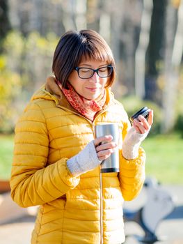 Happy wide smiling women in bright yellow jacket is holding thermos mug. Hot tea or other beverage on cool autumn day.