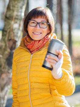Happy wide smiling women in bright yellow jacket is holding thermos mug. Hot tea or other beverage on cool autumn day.