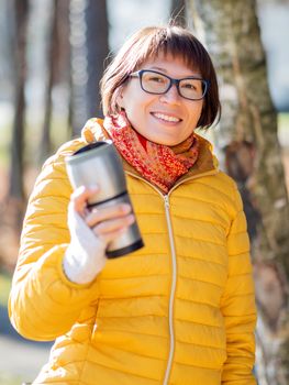 Happy wide smiling women in bright yellow jacket is holding thermos mug. Hot tea or other beverage on cool autumn day.