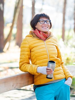 Happy wide smiling women in bright yellow jacket is holding thermos mug. Hot tea or other beverage on cool autumn day.