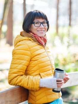 Happy wide smiling women in bright yellow jacket is holding thermos mug. Hot tea or other beverage on cool autumn day.