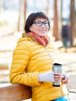 Happy wide smiling women in bright yellow jacket is holding thermos mug. Hot tea or other beverage on cool autumn day.