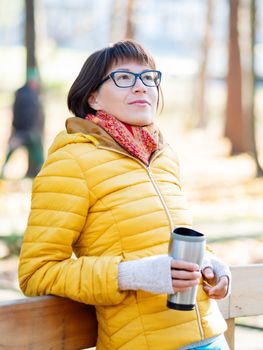 Happy wide smiling women in bright yellow jacket is holding thermos mug. Hot tea or other beverage on cool autumn day.