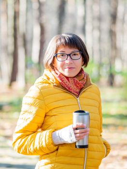 Happy wide smiling women in bright yellow jacket is holding thermos mug. Hot tea or other beverage on cool autumn day.