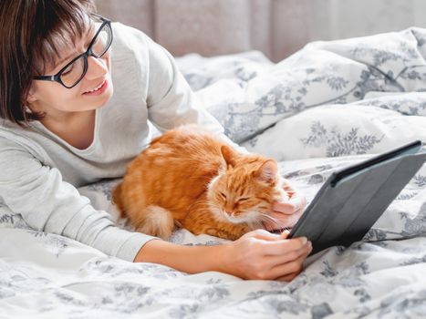 Cute ginger cat and woman in glasses are lying in bed. Woman is holding tablet and stroking her fluffy pet. Morning bedtime.