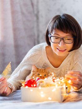 Curisous and cute ginger cat want to play with in light bulb garland. Fluffy pet and box with Christmas decorations. Cozy home before New Year.