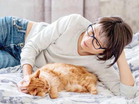 Cute ginger cat and woman in glasses are lying in bed. Woman is stroking her fluffy pet. Morning bedtime.