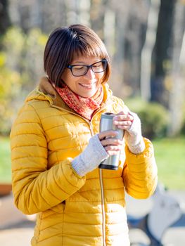 Happy wide smiling women in bright yellow jacket is holding thermos mug. Hot tea or other beverage on cool autumn day.