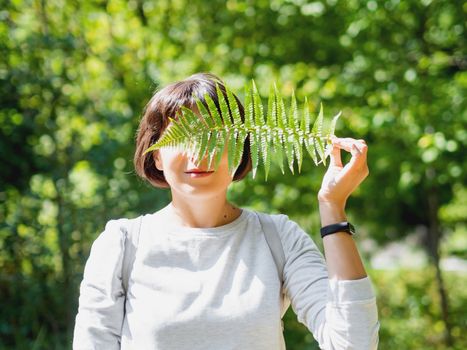 Young woman is hiding her eyes with fern leaf. Symbol of life, tranquility and unity with nature. Summer in forest.