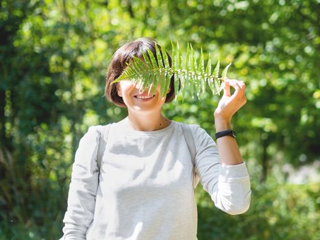 Young woman is hiding her eyes with fern leaf. Symbol of life, tranquility and unity with nature. Summer in forest.