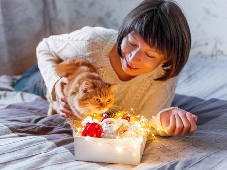 Curisous and cute ginger cat want to play with in light bulb garland. Fluffy pet and box with Christmas decorations. Cozy home before New Year.