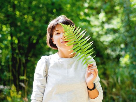 Young woman is hiding her eyes with fern leaf. Symbol of life, tranquility and unity with nature. Summer in forest.