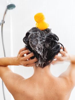 Naked woman with short hair takes a shower. Woman washes her hair with shampoo. Yellow rubber duck on girl's head in white bathroom.