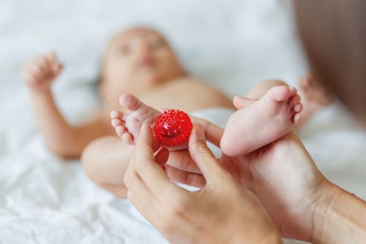 Mother holds newborn baby's feet. Tiny fingers and red massage ball in woman's hand. Cozy morning at home.