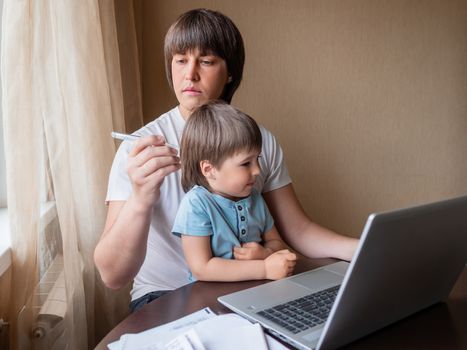 Self-isolation because of coronavirus outbreak. Man is remote working with his little son. Dad looks at the thermometer. Laptop on table.