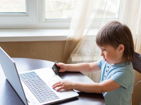 Curious toddler boy explores the laptop and presses buttons on computer keyboard. Kid holds credit card in hand. Puzzled child.