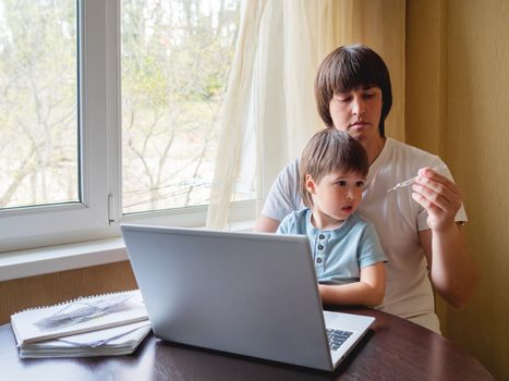 Self-isolation because of coronavirus outbreak. Man is remote working with his little son. Dad looks at the thermometer. Laptop on table.