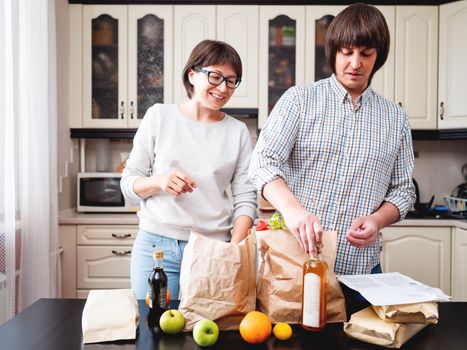 Young couple is sorting out purchases in the kitchen. Products in bags made of craft paper. Food delivery in conditions of quarantine because of coronavirus COVID19.