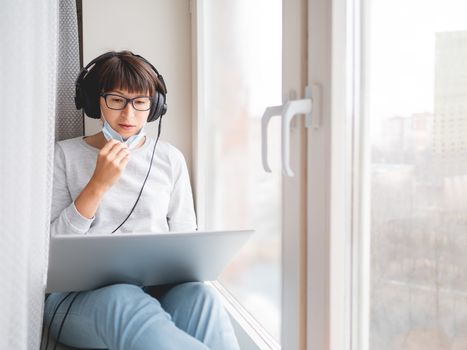 Woman in medical mask remote works from home. She sits on window sill with laptop on knees and headphones. Lockdown quarantine because of coronavirus COVID19. Self isolation at home.