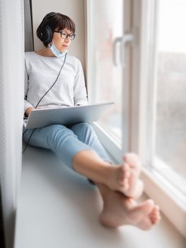 Woman in medical mask remote works from home. She sits on window sill with laptop on knees and headphones. Lockdown quarantine because of coronavirus COVID19. Self isolation at home.