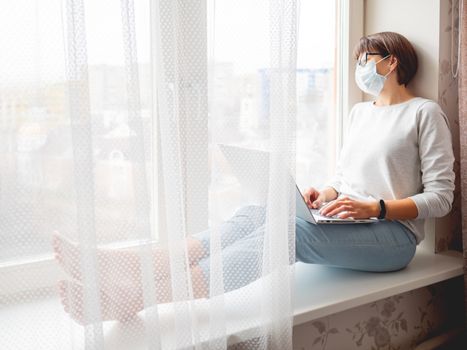 Woman in medical mask remote works from home. She sits on window sill with laptop on knees. Lockdown quarantine because of coronavirus COVID19. Self isolation at home.