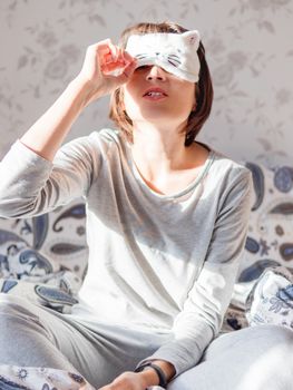 Young woman in grey pajama and sleeping mask in shape of cute sleeping cat face. She is just woke up and sit in bed. Early morning in cozy home.