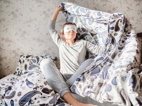 Young woman in grey pajama and sleeping mask in shape of cute sleeping cat face. She is just woke up and sit in bed. Early morning in cozy home.