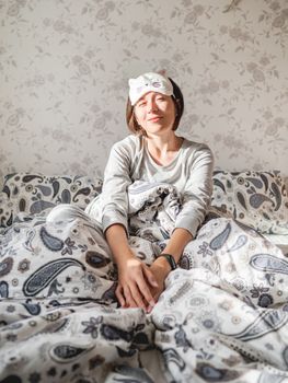 Young woman in grey pajama and sleeping mask in shape of cute sleeping cat face. She is just woke up and sit in bed. Early morning in cozy home.