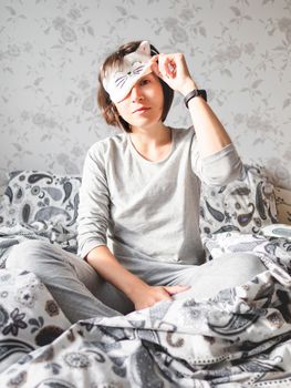 Young woman in grey pajama and sleeping mask in shape of cute sleeping cat face. She is just woke up and sit in bed. Early morning in cozy home.