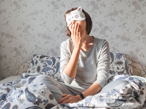 Young woman in grey pajama and sleeping mask in shape of cute sleeping cat face. She is just woke up and sit in bed. Early morning in cozy home.