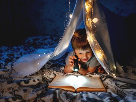 Little boy reads with pocket light. Toddler plays in tent made of linen sheet on bed. Cozy evening with favorite book.