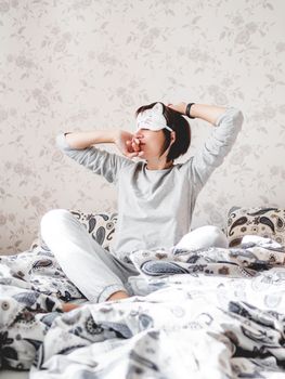 Young woman in grey pajama and sleeping mask in shape of cute sleeping cat face. She is just woke up and sit in bed. Early morning in cozy home.