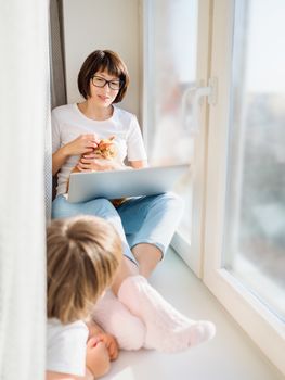 Woman in medical mask remote works from home. She sits on window sill with laptop and cute ginger cat on her knees. Lockdown quarantine because of coronavirus COVID19. Self isolation at home.