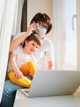 Woman in medical mask remote works from home. She sits on window sill with laptop and cute toddler boy on her knees. Lockdown quarantine because of coronavirus COVID19. Self isolation at home.