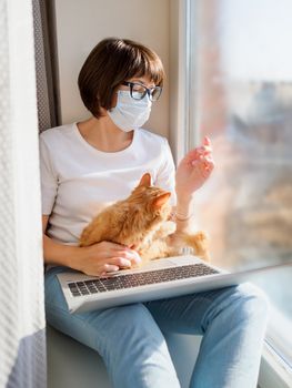 Woman in medical mask remote works from home. She sits on window sill with laptop and cute ginger cat on her knees. Lockdown quarantine because of coronavirus COVID19. Self isolation at home.