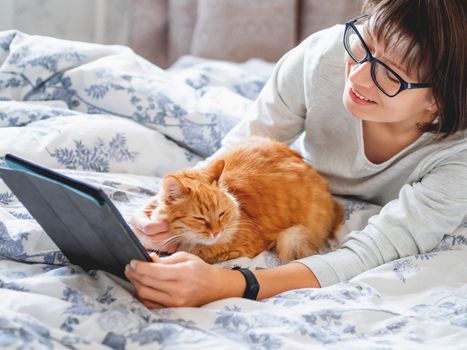 Cute ginger cat and woman in glasses are lying in bed. Woman is holding tablet and stroking her fluffy pet. Morning bedtime.