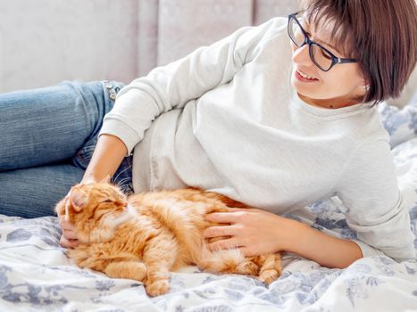 Cute ginger cat and woman in glasses are lying in bed. Woman is stroking her fluffy pet. Morning bedtime.