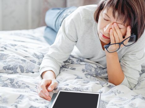 Woman in glasses are lying in bed with tablet. She is tired and rubs her eyes. Morning bedtime.