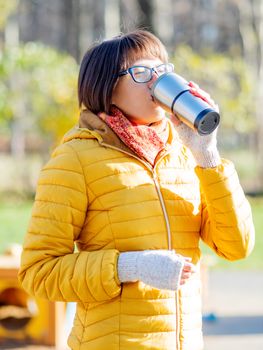 Happy wide smiling women in bright yellow jacket is holding thermos mug. Hot tea or other beverage on cool autumn day.