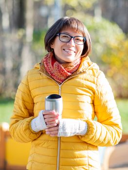 Happy wide smiling women in bright yellow jacket is holding thermos mug. Hot tea or other beverage on cool autumn day.