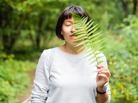 Young woman is hiding her eye with fern leaf. Symbol of life, tranquility and unity with nature. Summer in forest.