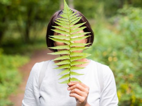 Young woman is hiding her eyes with fern leaf. Symbol of life, tranquility and unity with nature. Summer in forest.