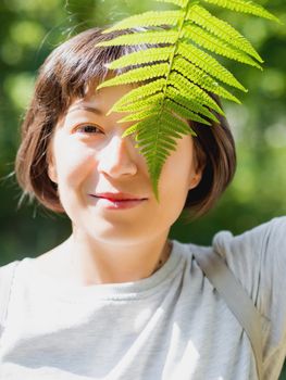 Young woman is hiding her eye with fern leaf. Symbol of life, tranquility and unity with nature. Summer in forest.