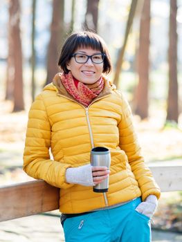 Happy wide smiling women in bright yellow jacketis holding thermos mug. Hot tea or other beverage on cool autumn day.