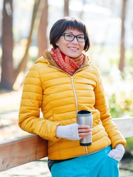 Happy wide smiling women in bright yellow jacket is holding thermos mug. Hot tea or other beverage on cool autumn day.